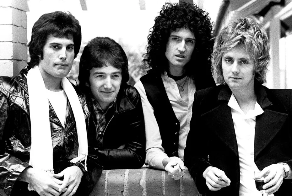 Black and white photo of Queen band members posing outdoors with casual 1970s style clothing and hairstyles.