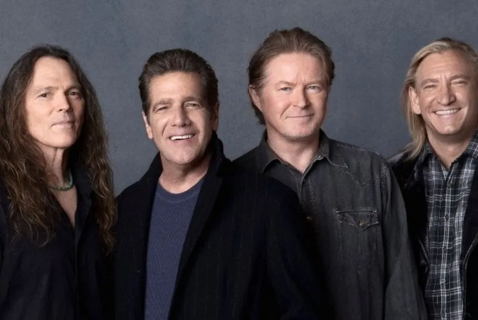 The Eagles band members posing together, smiling and dressed in casual dark clothing against a gray backdrop.