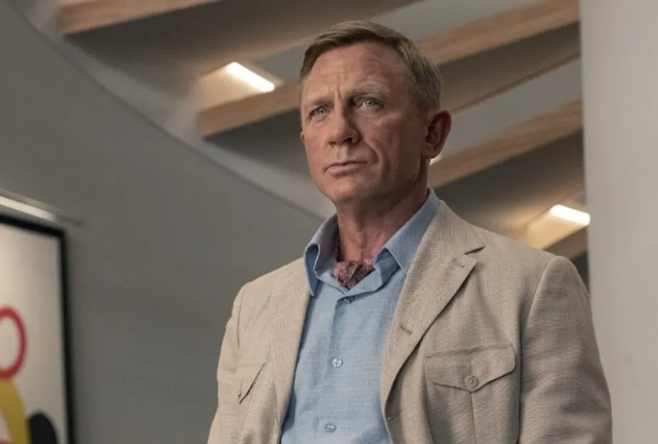 Daniel Craig as James Bond wearing a beige jacket and blue shirt in a modern indoor setting.