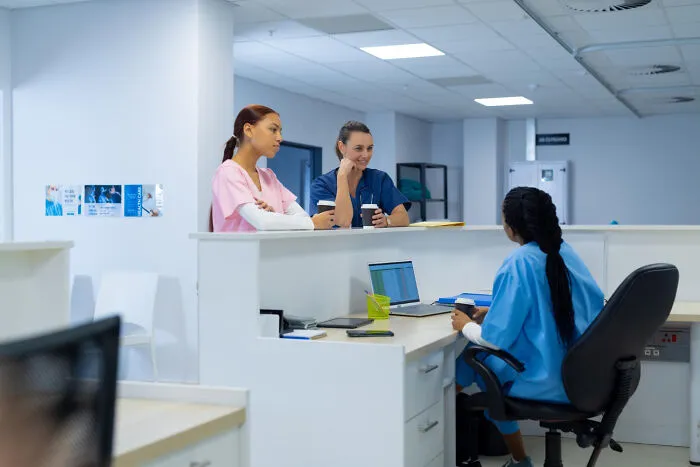 Three healthcare workers in scrubs having a discussion in a modern hospital nursing station environment