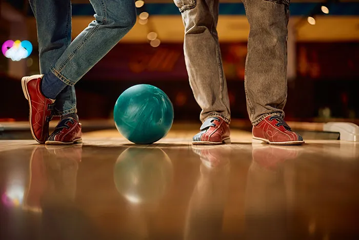 Two people wearing bowling shoes standing near a teal bowling ball, illustrating first dates with character-building trauma.
