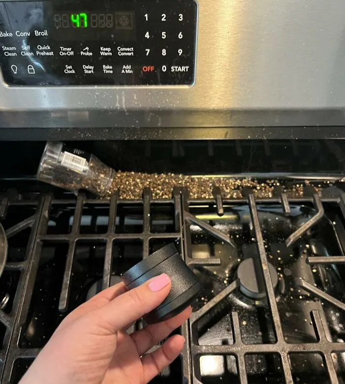 Close-up of spilled pepper on a stove burner with a hand holding a black lid, illustrating an odd and unexplained kitchen mishap.