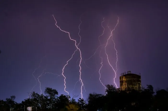Multiple lightning bolts striking near a silhouetted structure and trees during a nighttime thunderstorm capturing fake facts about nature.