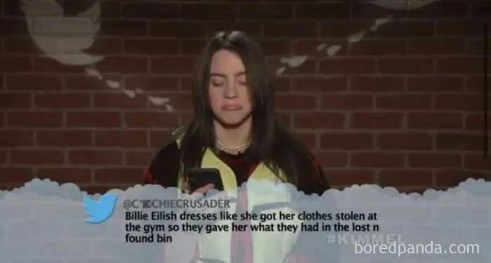 Billie Eilish reacting to a mean tweet that hits hard, showcasing a painful burn, in front of a brick wall.