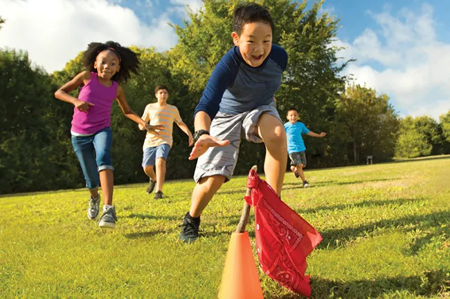 Children playing capture the flag outdoors, running on grass with a red flag tied to an orange cone on a sunny day.