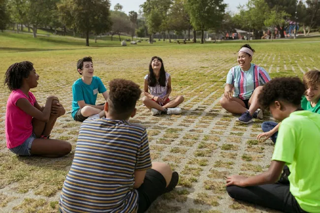 Children sitting in a circle outside playing Duck Duck Goose on grass, enjoying a classic childhood game for adults.