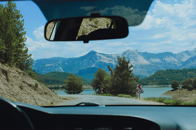 View from inside a car on a last-minute road trip with mountains, trees, and a lake under a blue sky ahead.