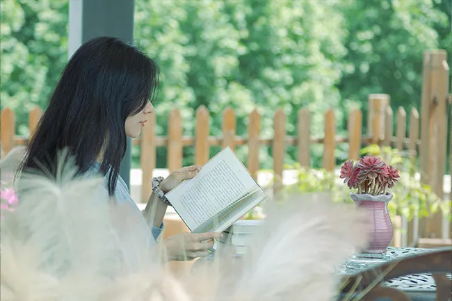 Woman enjoying a staycation with books outdoors near a wooden fence and potted flowers on a sunny day
