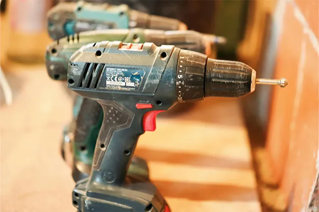 Power drill with a drill bit attached, placed on a wooden surface along with other drills in a workshop setting.