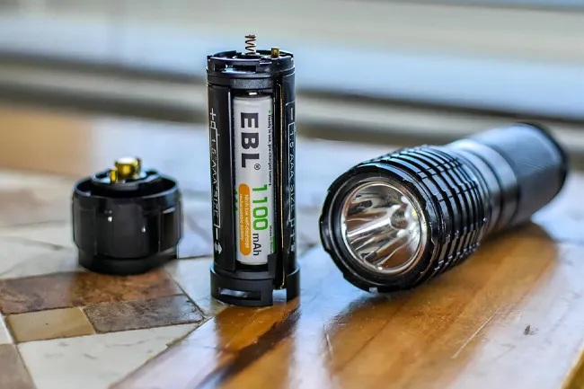 Flashlight batteries inside a battery holder next to a black flashlight resting on a wooden surface at home.