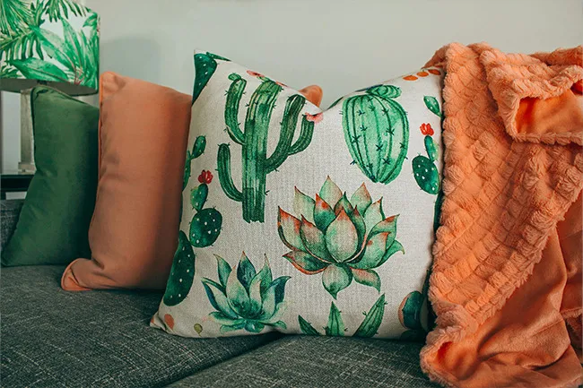 Decorative pillows with cactus and succulent designs on a couch surrounded by solid color pillows and an orange throw blanket.