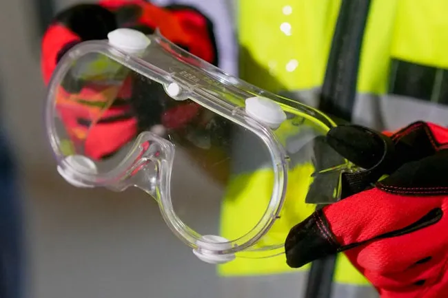 Clear safety goggles held by a person wearing red gloves and a yellow safety vest, ready for protection.