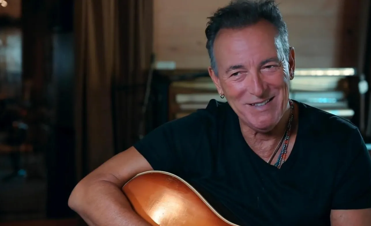 Famous musician from '80s and '90s music icons smiling and holding an acoustic guitar in a cozy indoor setting.