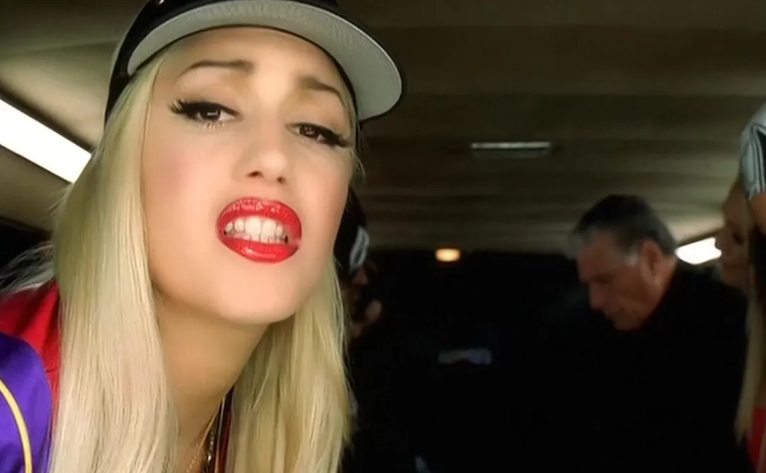 Close-up of a blonde music icon from the '80s and '90s wearing a cap and red lipstick in a dimly lit setting.