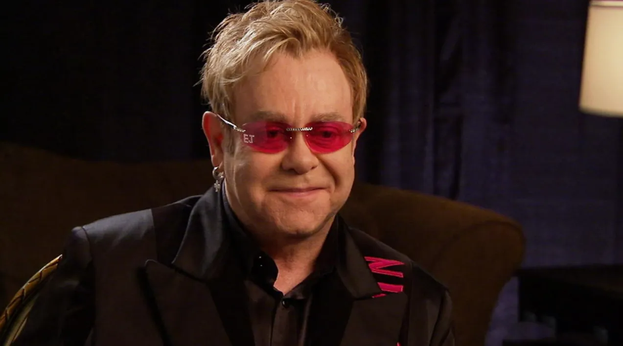 Male 80s and 90s music icon wearing red-tinted glasses and a black jacket, smiling in an indoor setting.