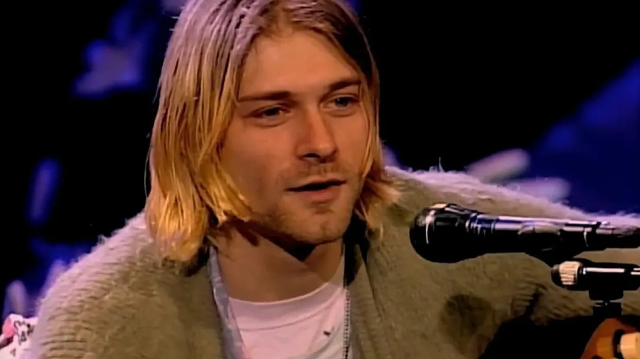 Male '80s and '90s music icon singing into a microphone with shoulder-length blonde hair and casual attire.