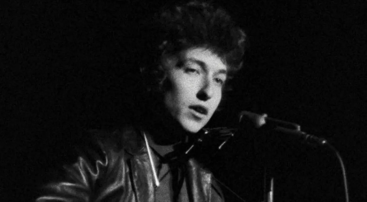 Black and white image of an '80s music icon performing on stage with a microphone, wearing a leather jacket.