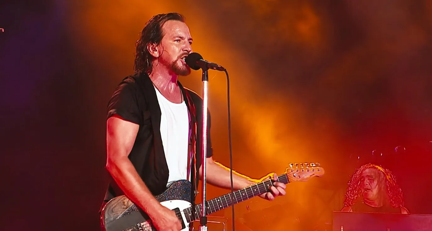 Well-known '80s and '90s music icon singing and playing guitar on stage with vibrant orange and red concert lighting.