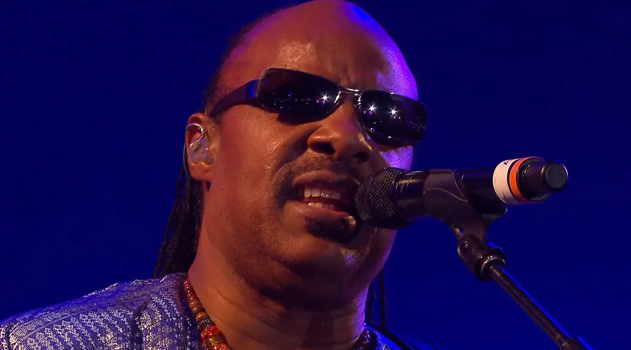 Famous '80s and '90s music icon singing on stage with sunglasses and colorful beaded necklace during live performance.