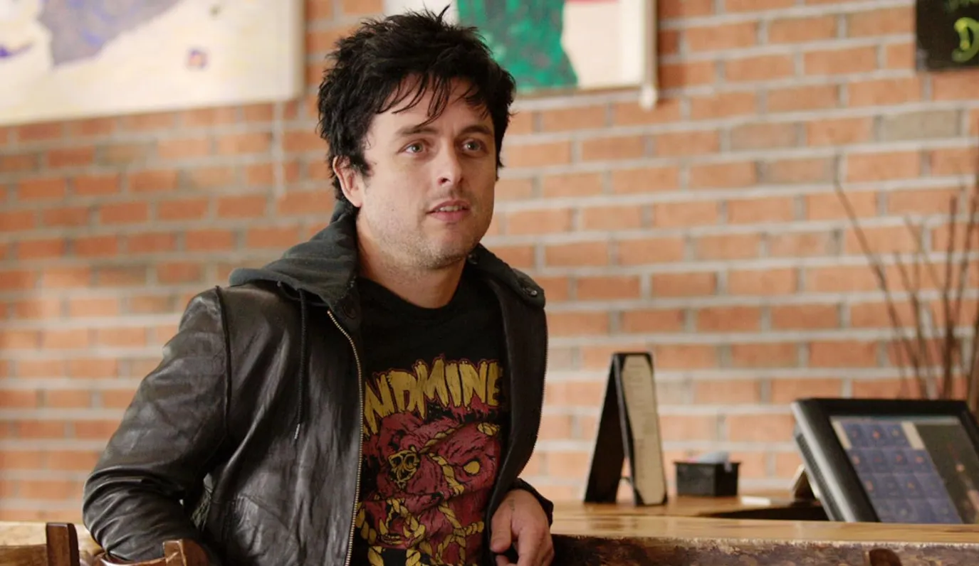 Man in a leather jacket and graphic tee sitting in a cafe with brick wall background name the 80s and 90s music icons