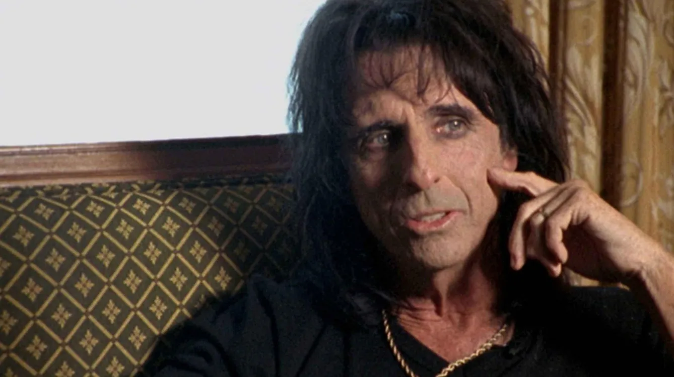 Famous '80s and '90s music icon sitting on patterned couch, wearing a black shirt and gold chain, thoughtful expression.