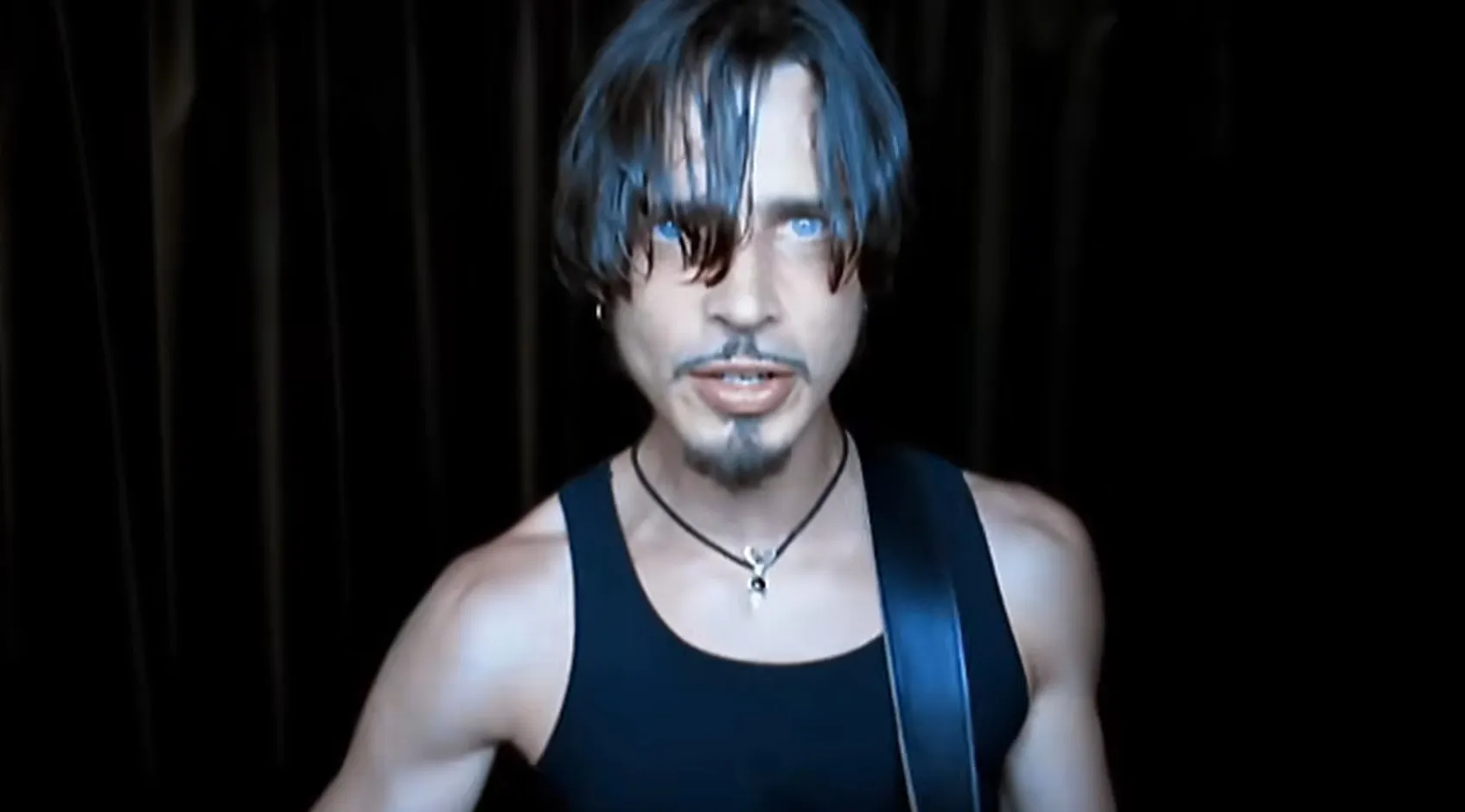 Music icon from the 80s and 90s wearing a black tank top and necklace performing against a dark background.