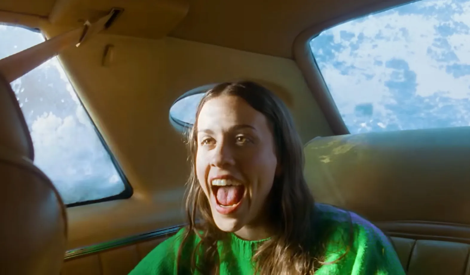 Young music icon from the 80s and 90s singing excitedly inside a vintage car with green sweater and open mouth.