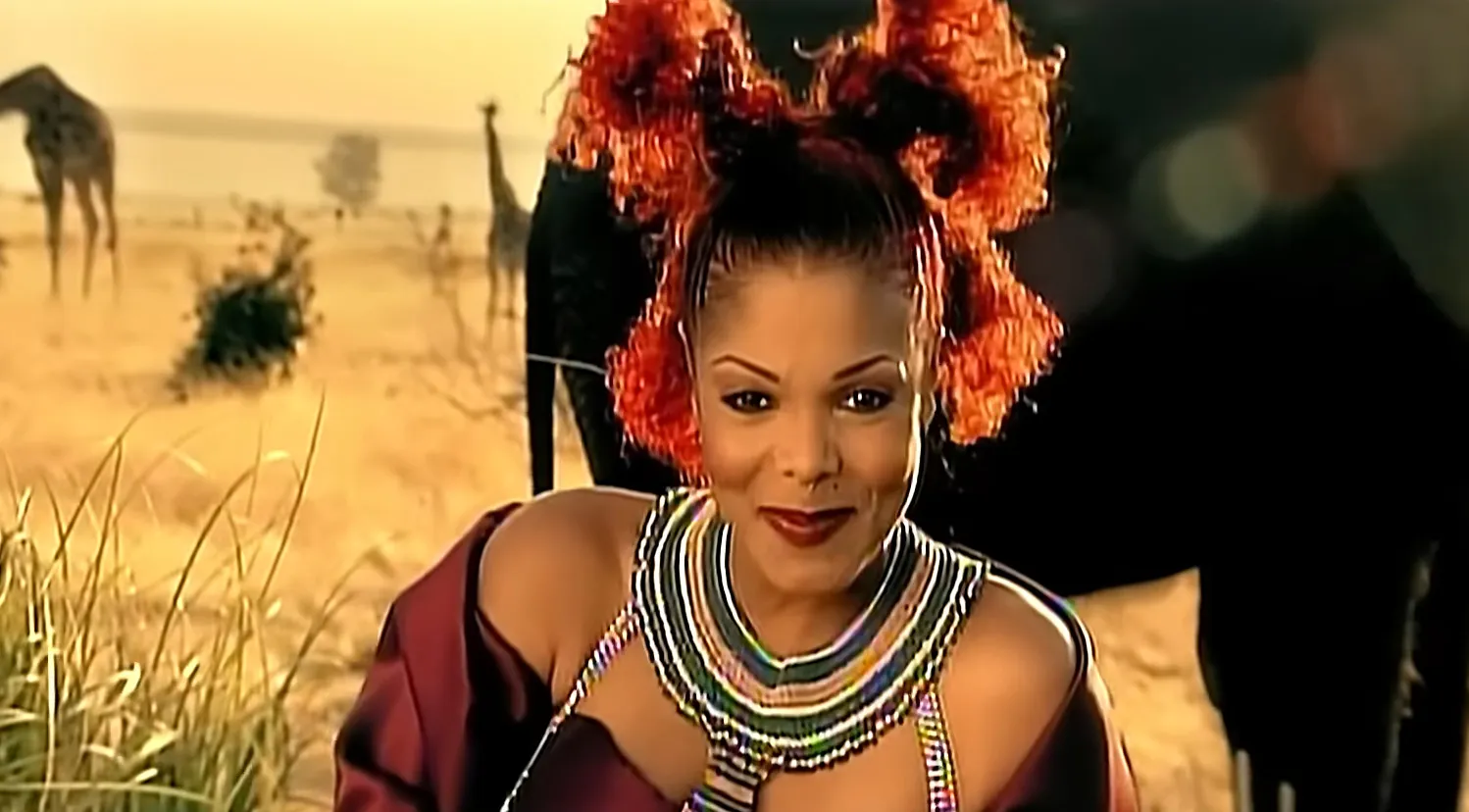 80s and 90s music icon with vibrant red hair and beaded necklace smiling in a savanna setting with giraffes in the background
