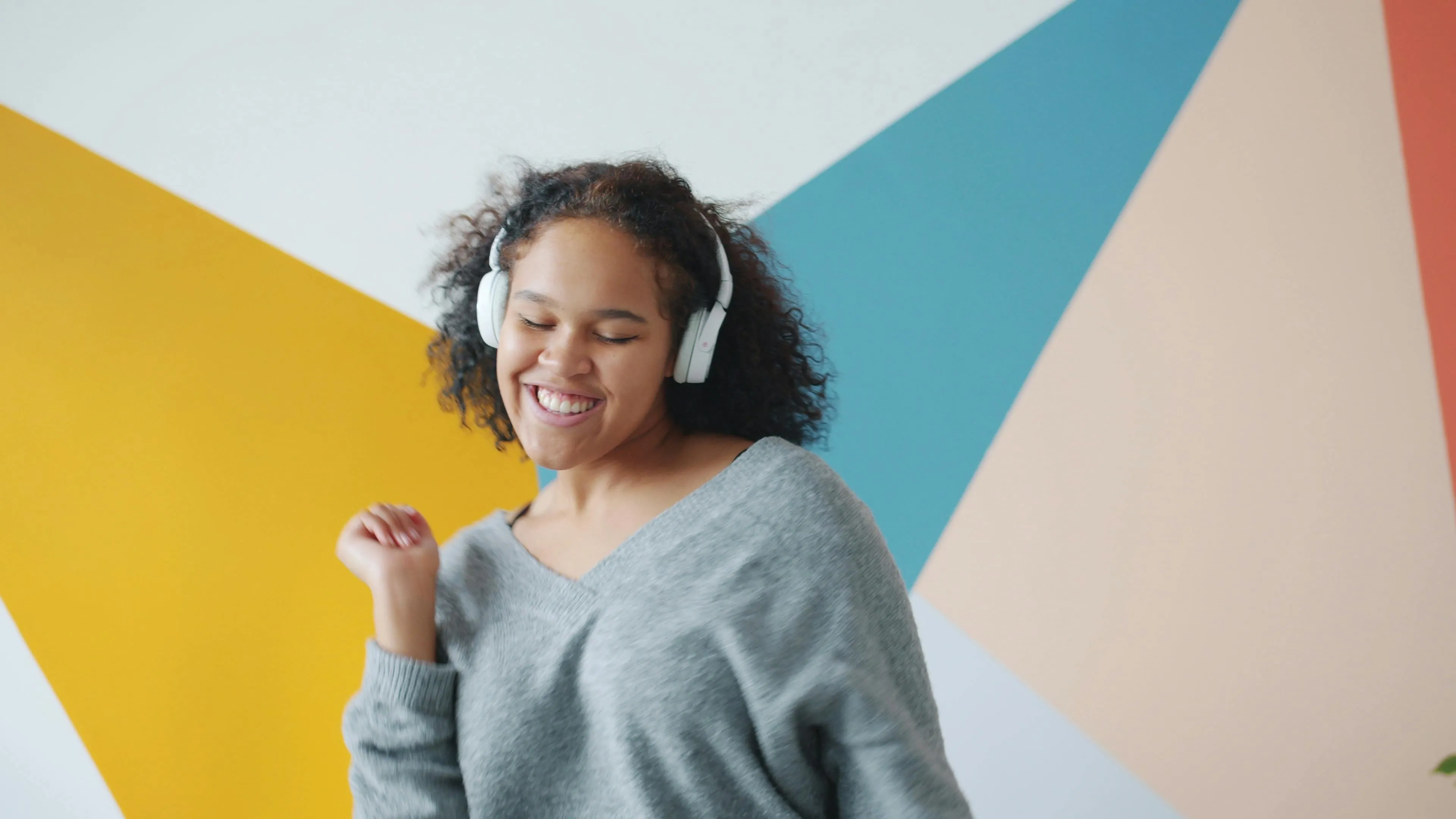 Young woman wearing headphones, smiling and dancing against a colorful geometric wall, enjoying a pub quiz music challenge.