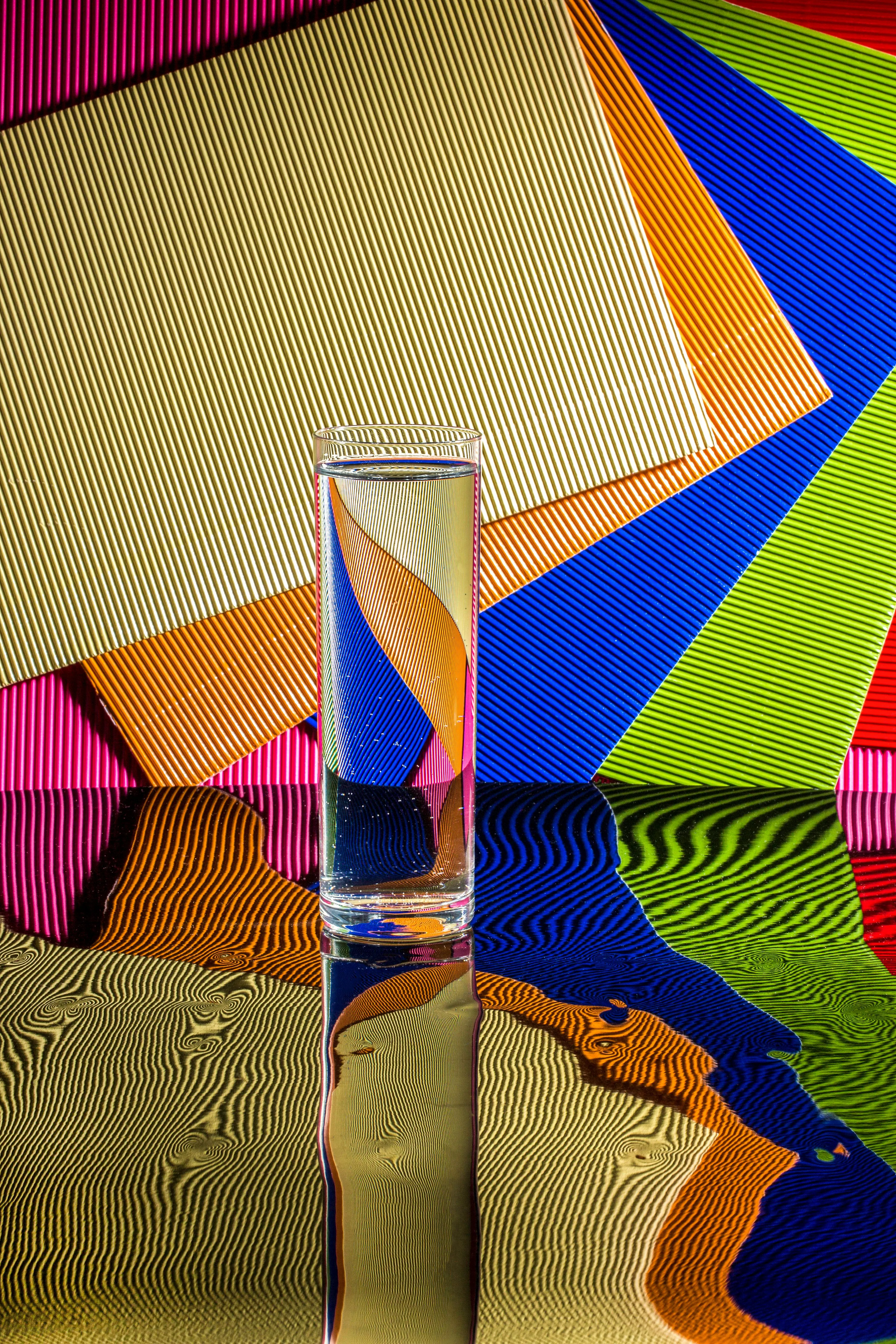Glass of water bending light rays with colorful striped background demonstrating refraction phenomenon.