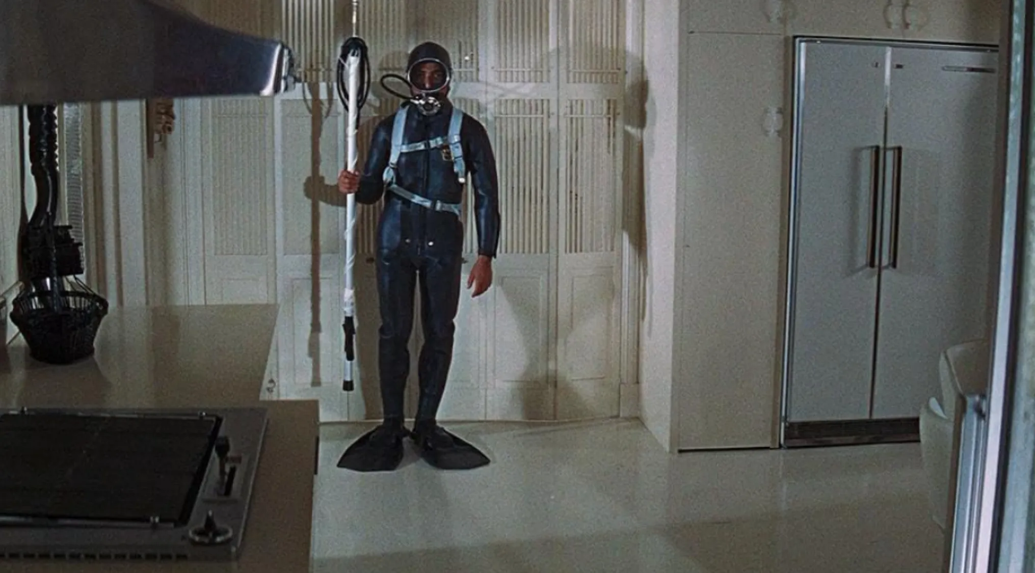 Man in a vintage diving suit holding equipment in a 60s movie scene inside a modern kitchen setting.