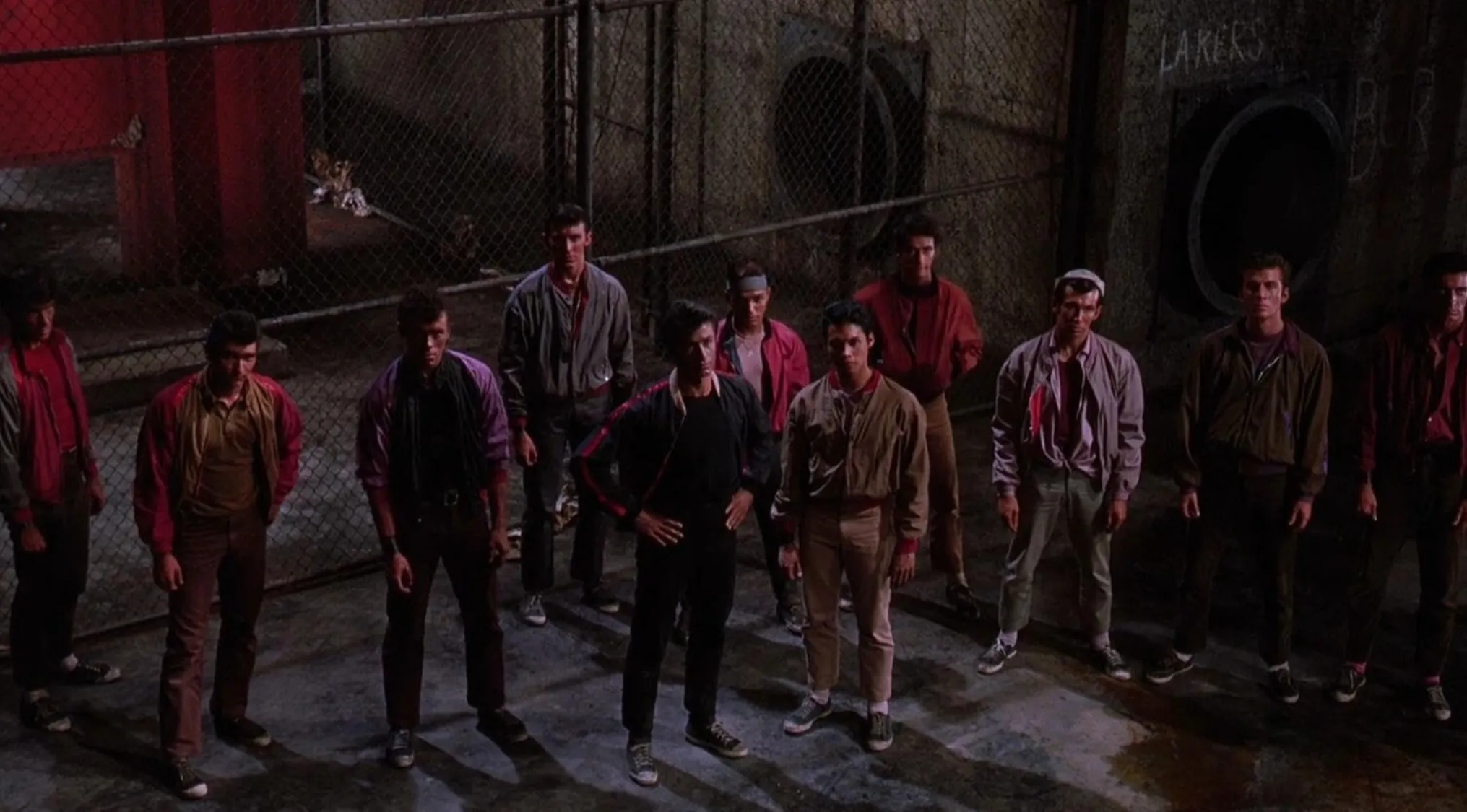 60s movie scenes showing a tense group of young men standing in a dimly lit urban alleyway near a chain-link fence