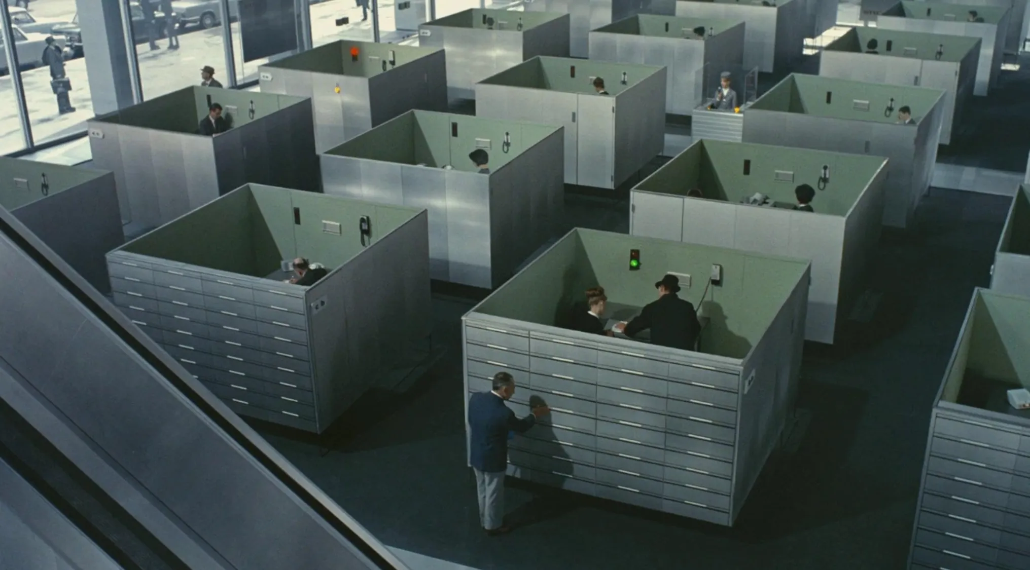 1960s movie scene showing large office cubicles with workers inside in a vintage corporate setting.