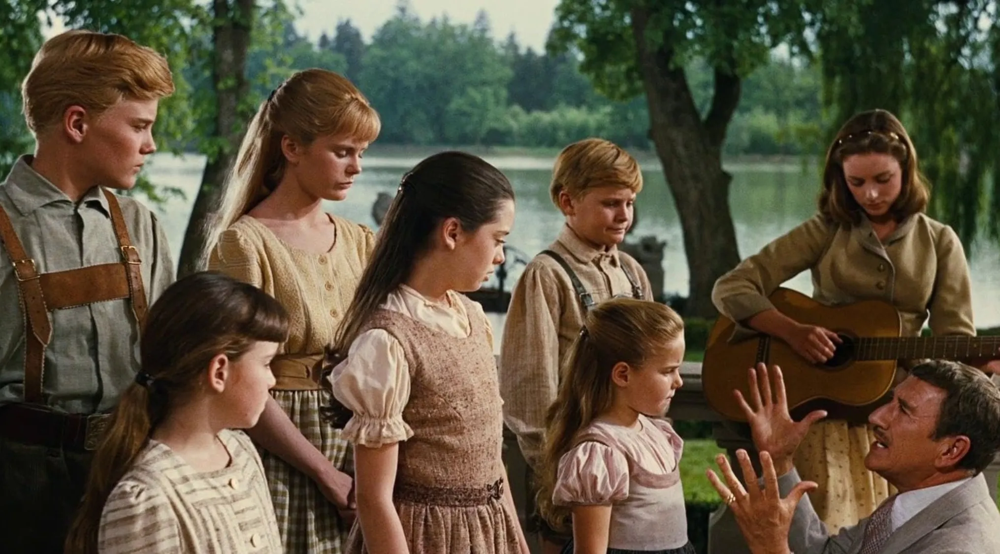 Children dressed in 60s style gathered outdoors while a man speaks and a woman plays guitar in a movie scene.