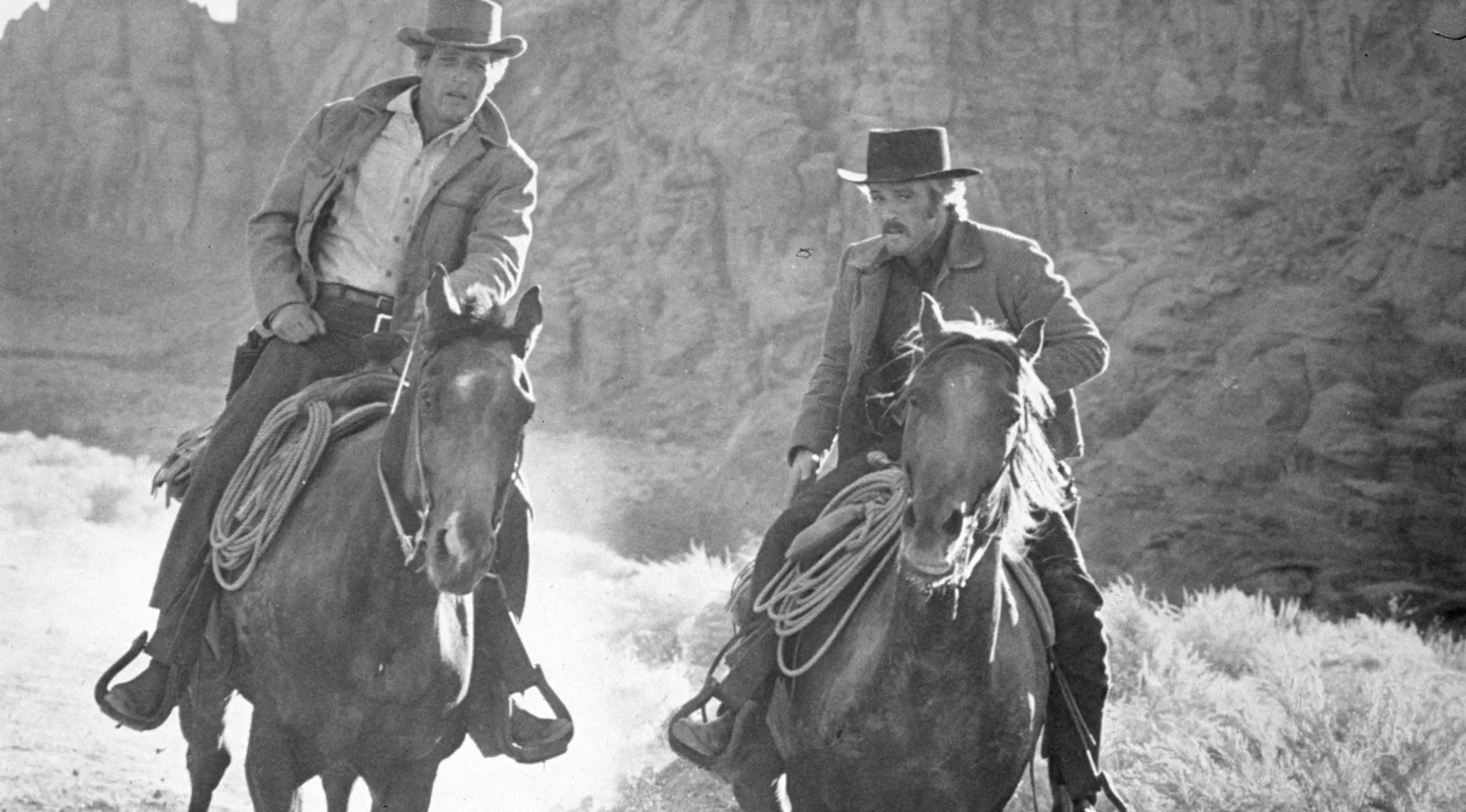 Two cowboys riding horses through a rocky desert landscape in a 60s movie scene.