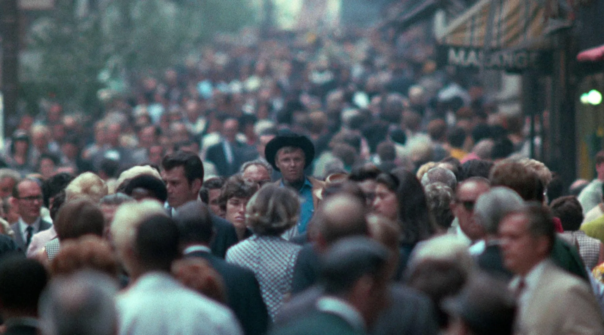 Crowded city street scene from 60s movie with a man in a cowboy hat walking among pedestrians and vintage storefronts.