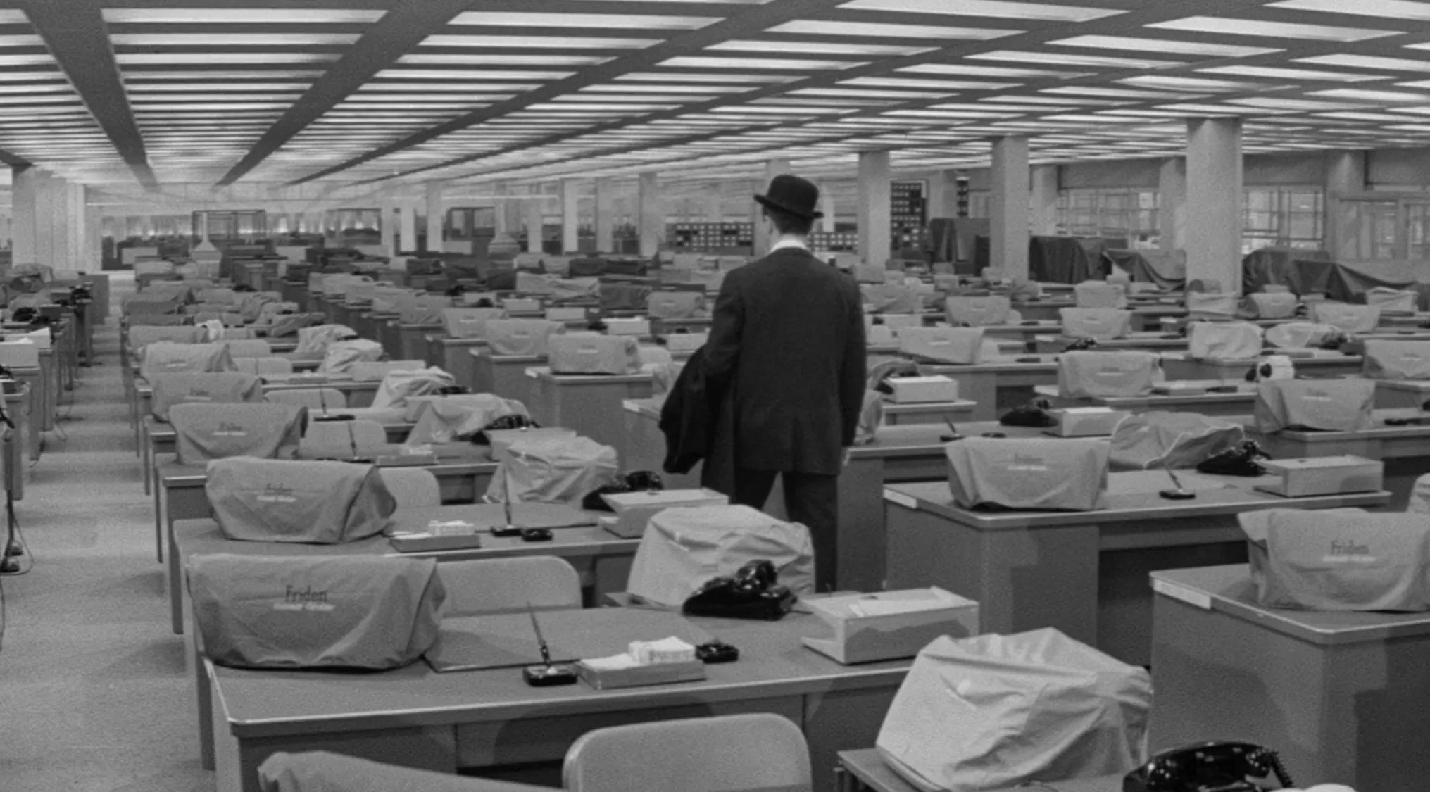 Man in a bowler hat stands in a vast 1960s movie office scene filled with covered typewriters and desks in black and white.