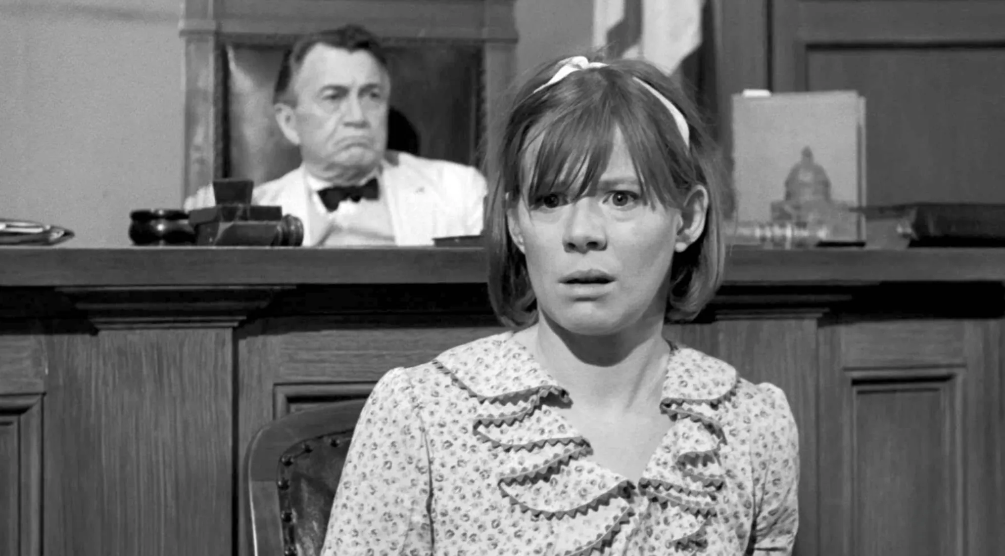 Black and white 60s movie scene showing a distressed woman in court with a judge seated behind her.