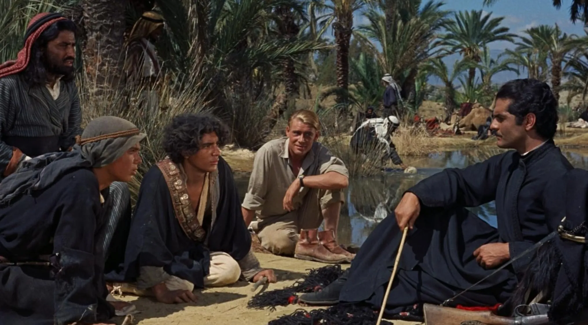 1960s movie scene with men in desert setting, palm trees, and traditional Middle Eastern attire by a water pool.