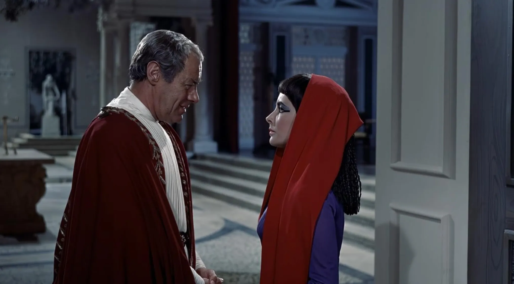 A scene from a 60s movie showing a man in a red cape speaking to a woman with dramatic black eye makeup and a red hood.