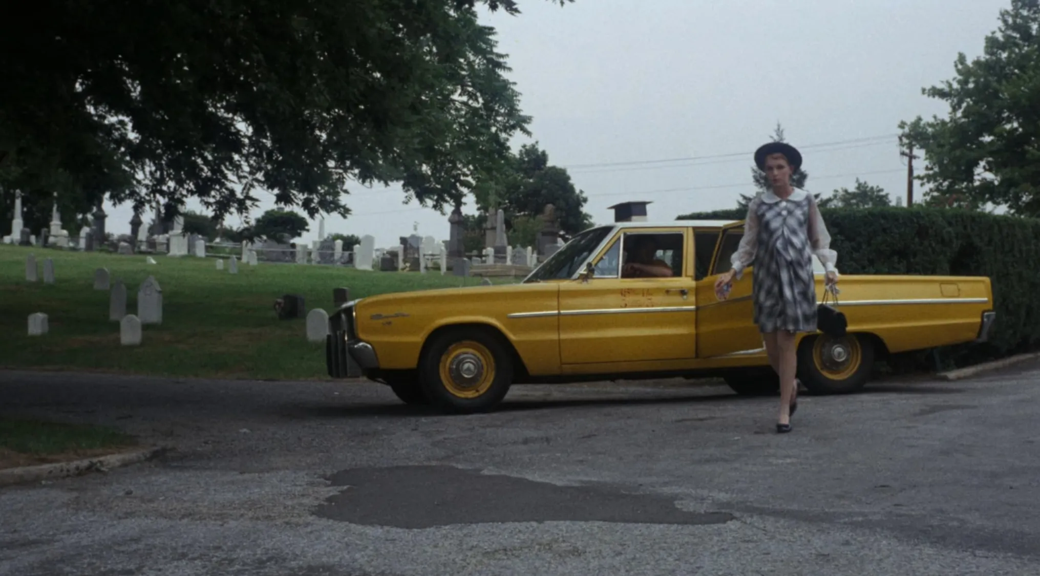 Woman in 60s movie scene walking near yellow vintage car in a cemetery setting for name the 60s film challenge
