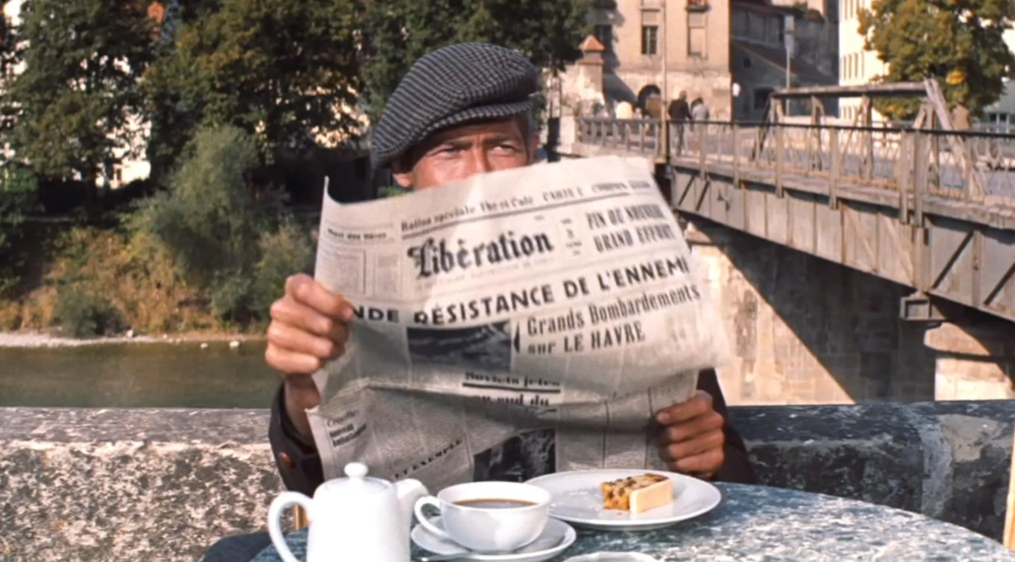 Man reading a newspaper at an outdoor table with coffee and cake in a classic 60s movie scene by a bridge.