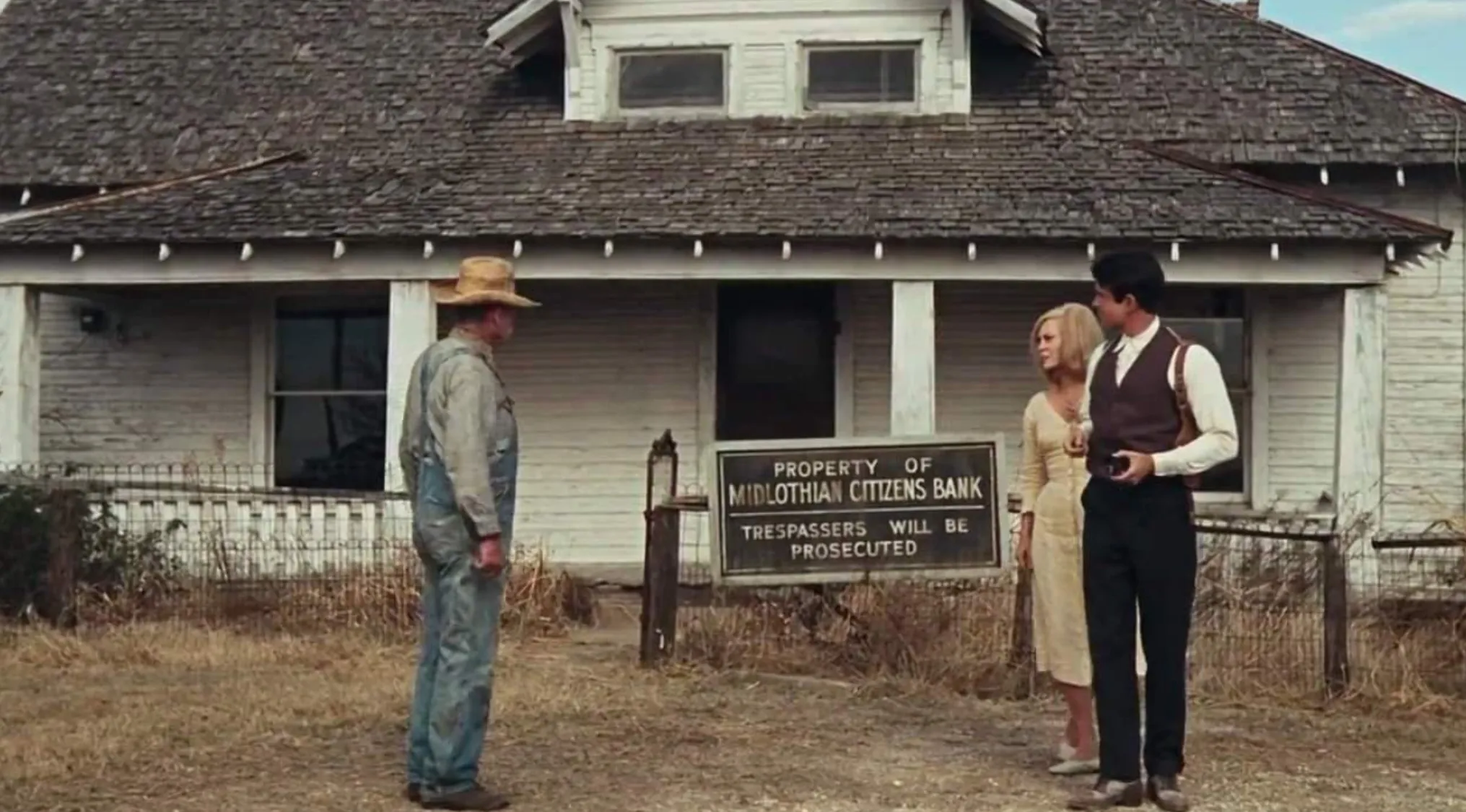 60s movie scene with three people standing near a worn fence and a house with a Midlothian Citizens Bank sign.