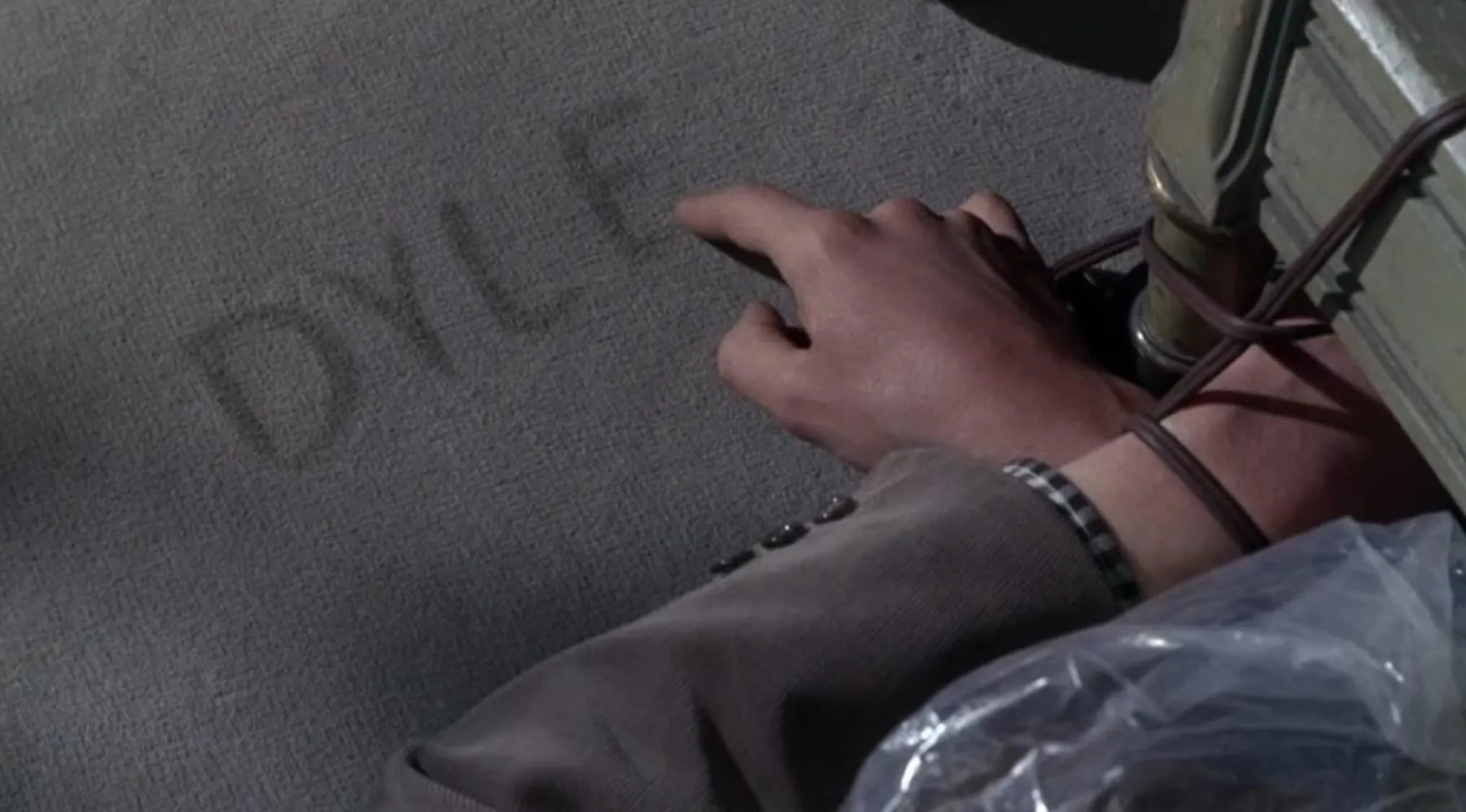 Hand tied to a bomb with a timer, pointing at a message on the carpet in a 60s movie scene.