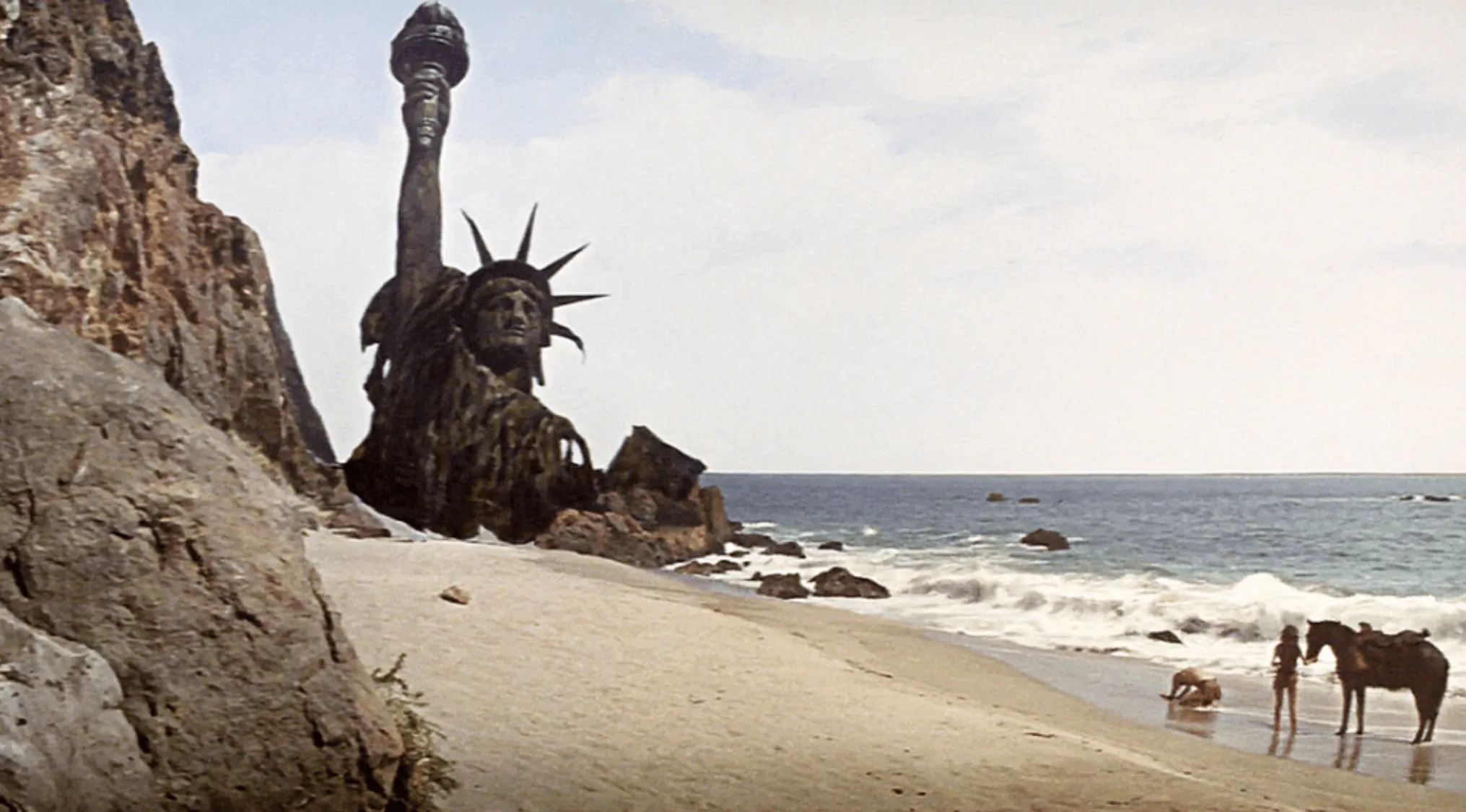 Scene from a 60s movie showing a ruined Statue of Liberty on a deserted beach with two people and a horse.