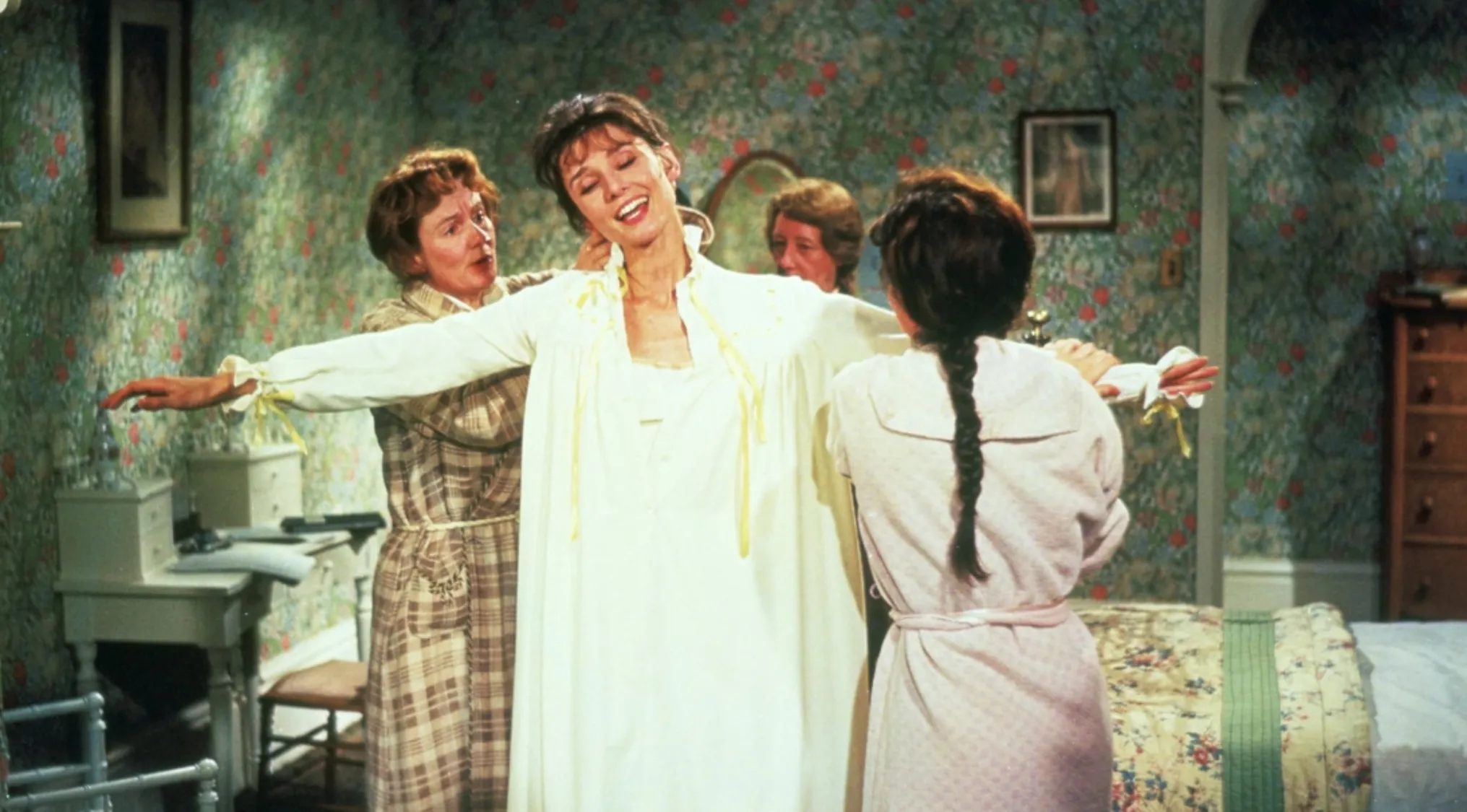 Women helping another woman into a white dress in a floral wallpapered bedroom from a 60s movie scene.