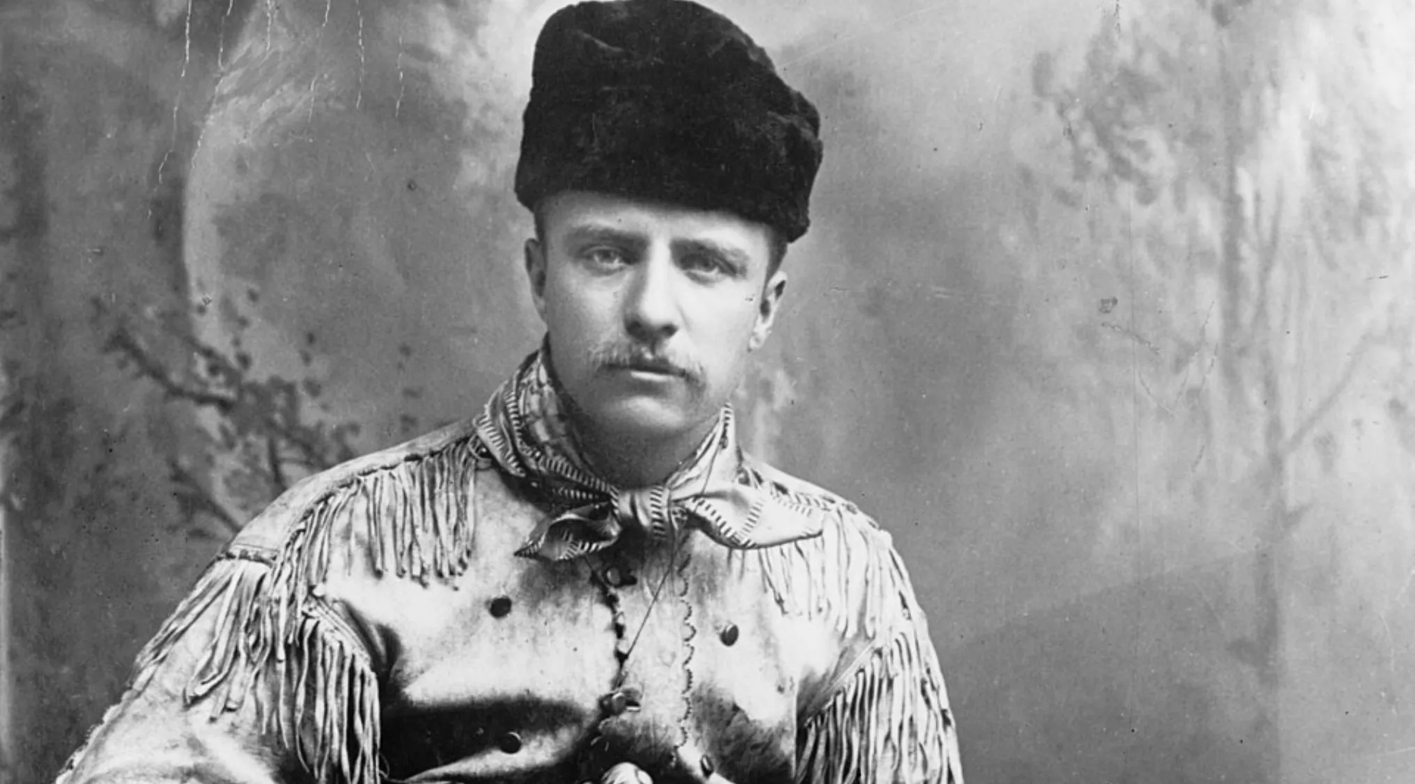 Black and white photo of famous president Theodore Roosevelt in a fringed jacket and fur hat looking at the camera.