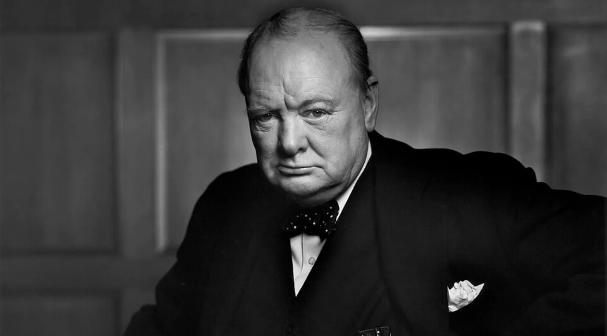 Black and white portrait of Winston Churchill wearing a bow tie and suit, posing seriously against a wooden background.