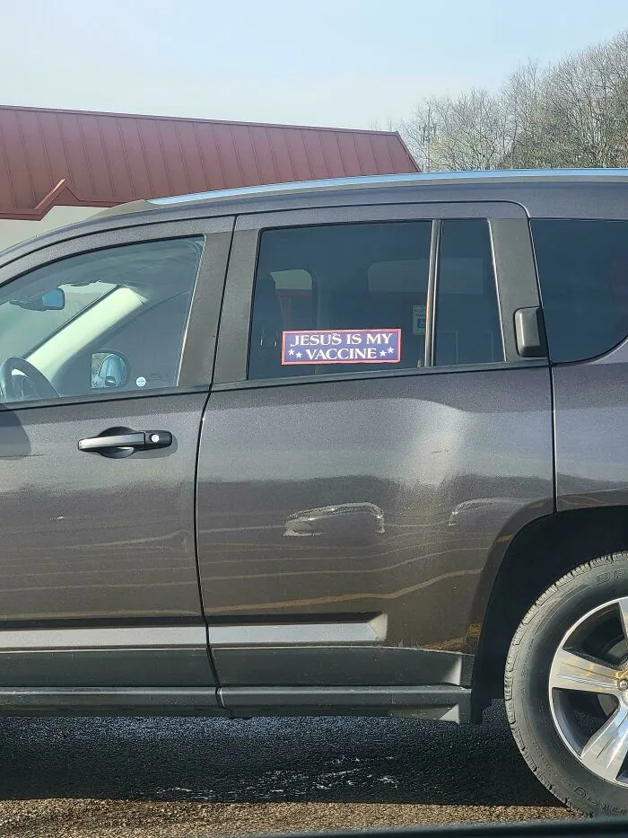 A car with an unfiltered bumper sticker on the window that says, JESUS IS MY VACCINE.