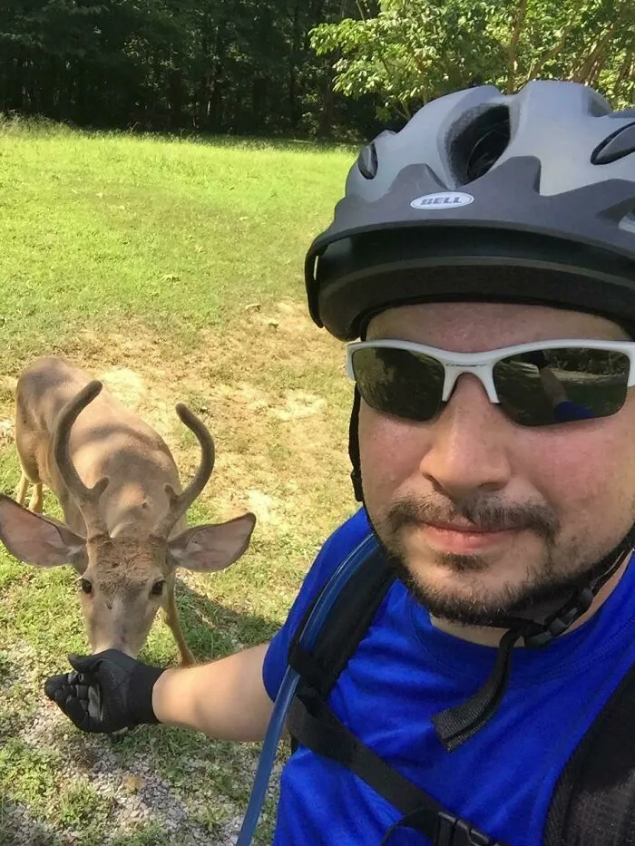 Man in a bike helmet and sunglasses having a wild and wholesome animal encounter with a young deer in a grassy area.